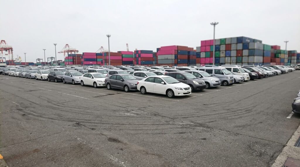 輸出を待つ中古車が並ぶ港のヤードと、奥に積まれた輸送用コンテナ