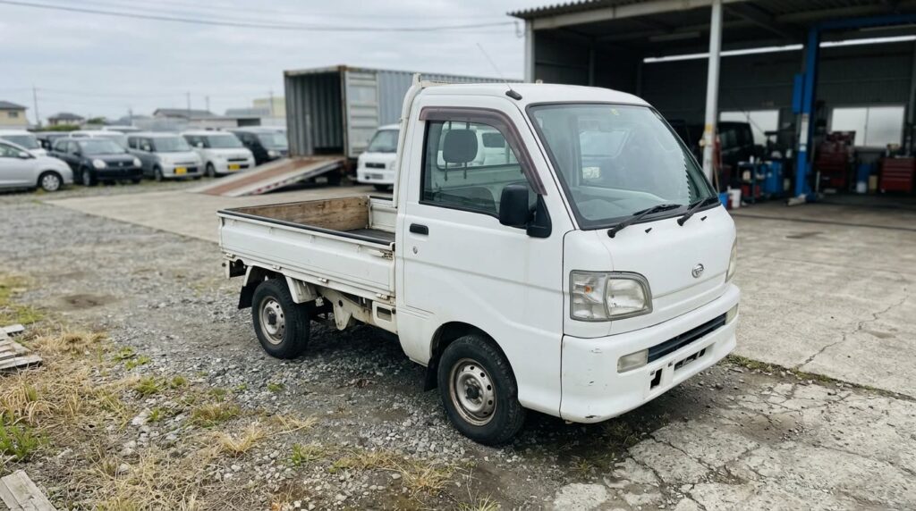 屋外のヤードに1台だけ置かれた荷台付きの小型商用トラックを斜め前から写した様子