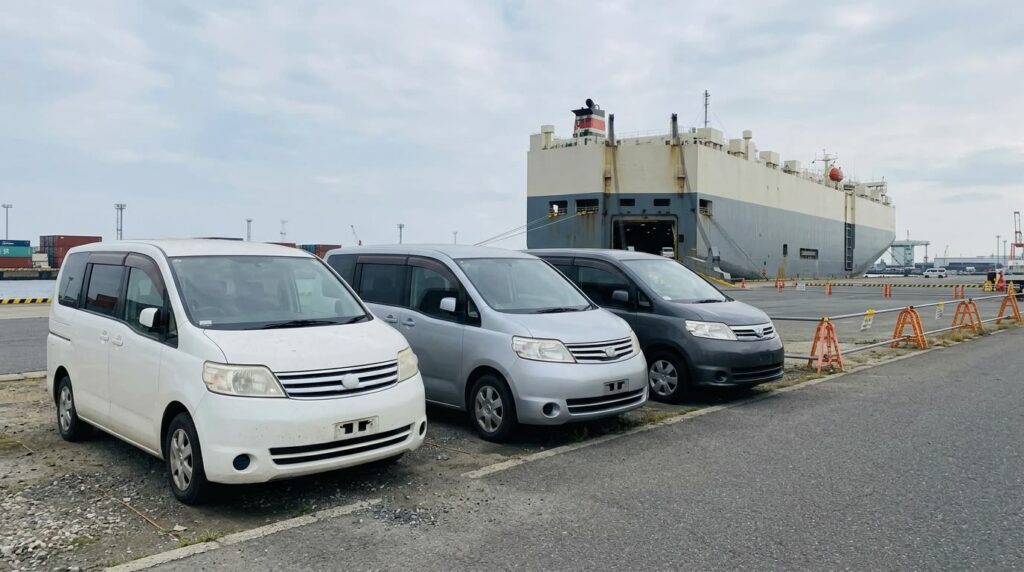 岸壁に並ぶ数台の中古ミニバンと自動車運搬船